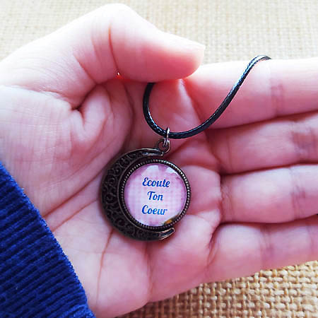 photographie d'une main qui tient un collier au pendentif eu forme de lune et au message inspirant Ecoute ton Coeur